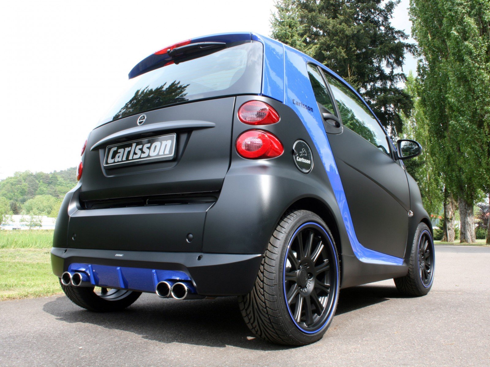A sleek black and blue Smart vehicle sits parked on a tree-lined road, showcasing its sporty design. This HD PC desktop wallpaper highlights modern automotive aesthetics.