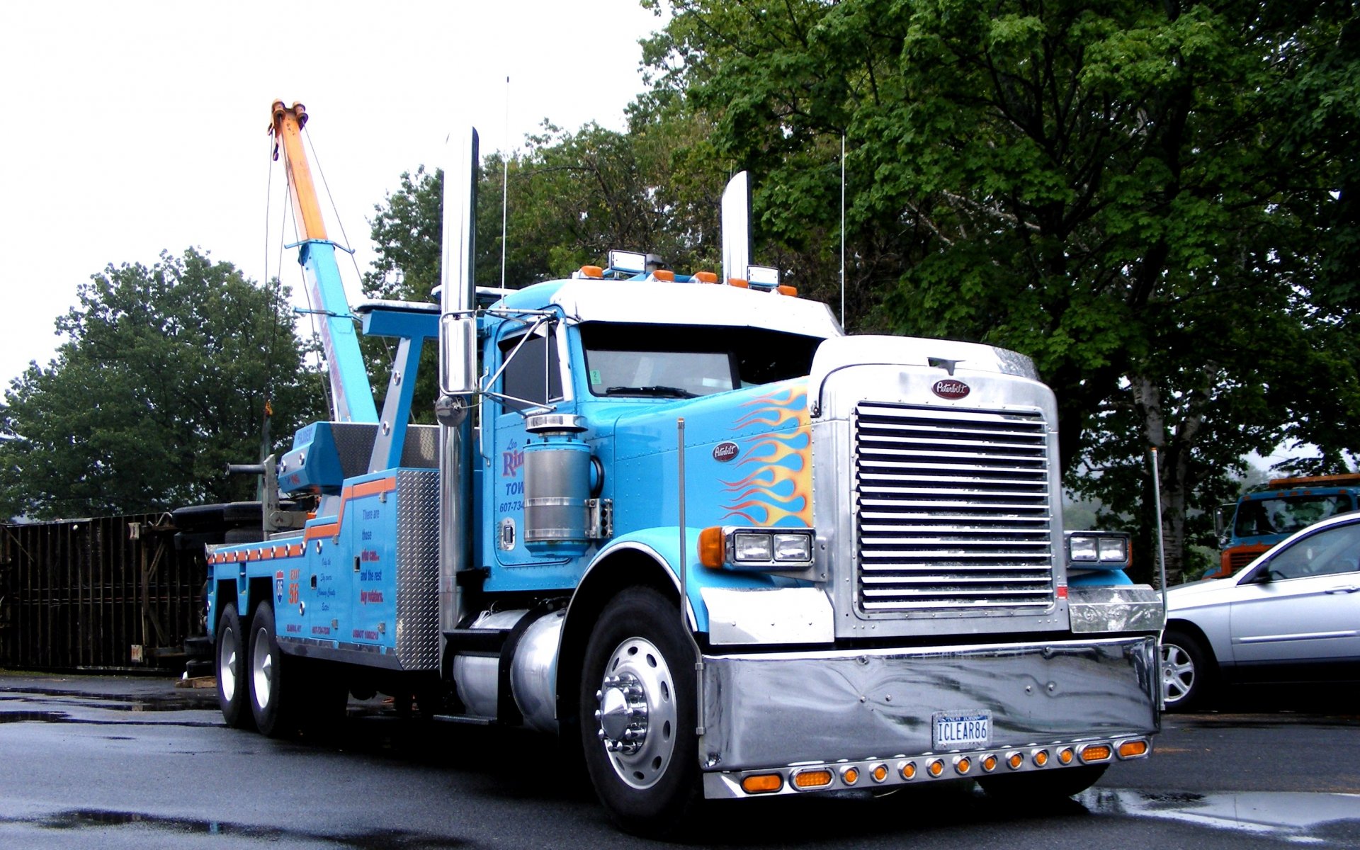 HD PC desktop wallpaper featuring a blue Peterbilt tow truck with flame decals, parked on a wet street with trees in the background.
