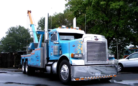 HD PC desktop wallpaper featuring a blue Peterbilt tow truck with flame decals, parked on a wet street with trees in the background.