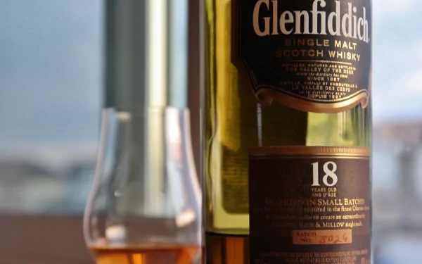 A close-up of a Glenfiddich 18-year-old single malt Scotch whisky bottle next to a filled glass, set against a blurred background, showcasing the rich colors and textures of whisky.