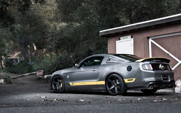 HD PC desktop wallpaper/background: a gray Ford Mustang GT with a yellow stripe parked beside a rustic garage under oak trees.