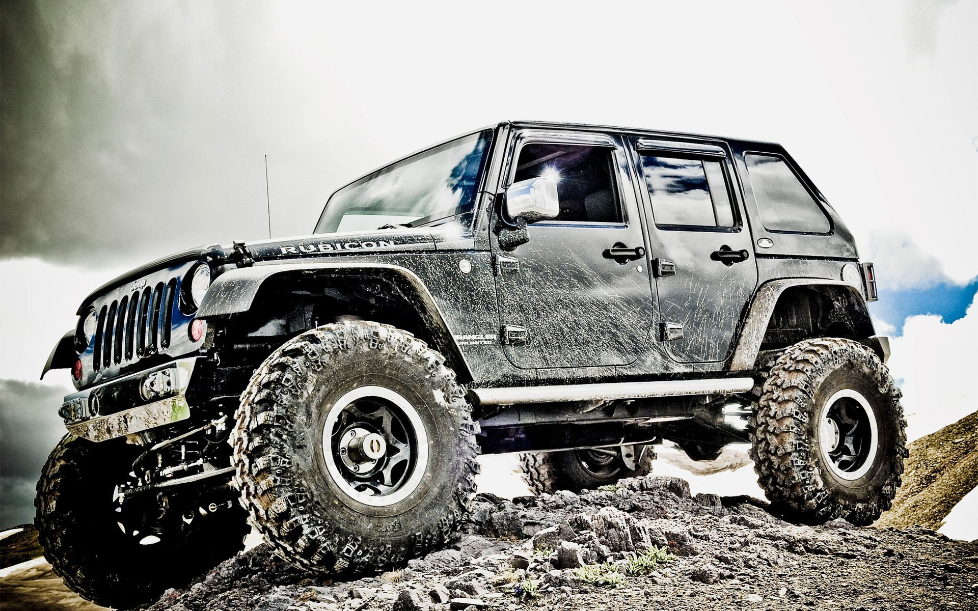 HD PC desktop wallpaper featuring a rugged Jeep with large off-road tires, positioned on rocky terrain under a bright, cloudy sky.