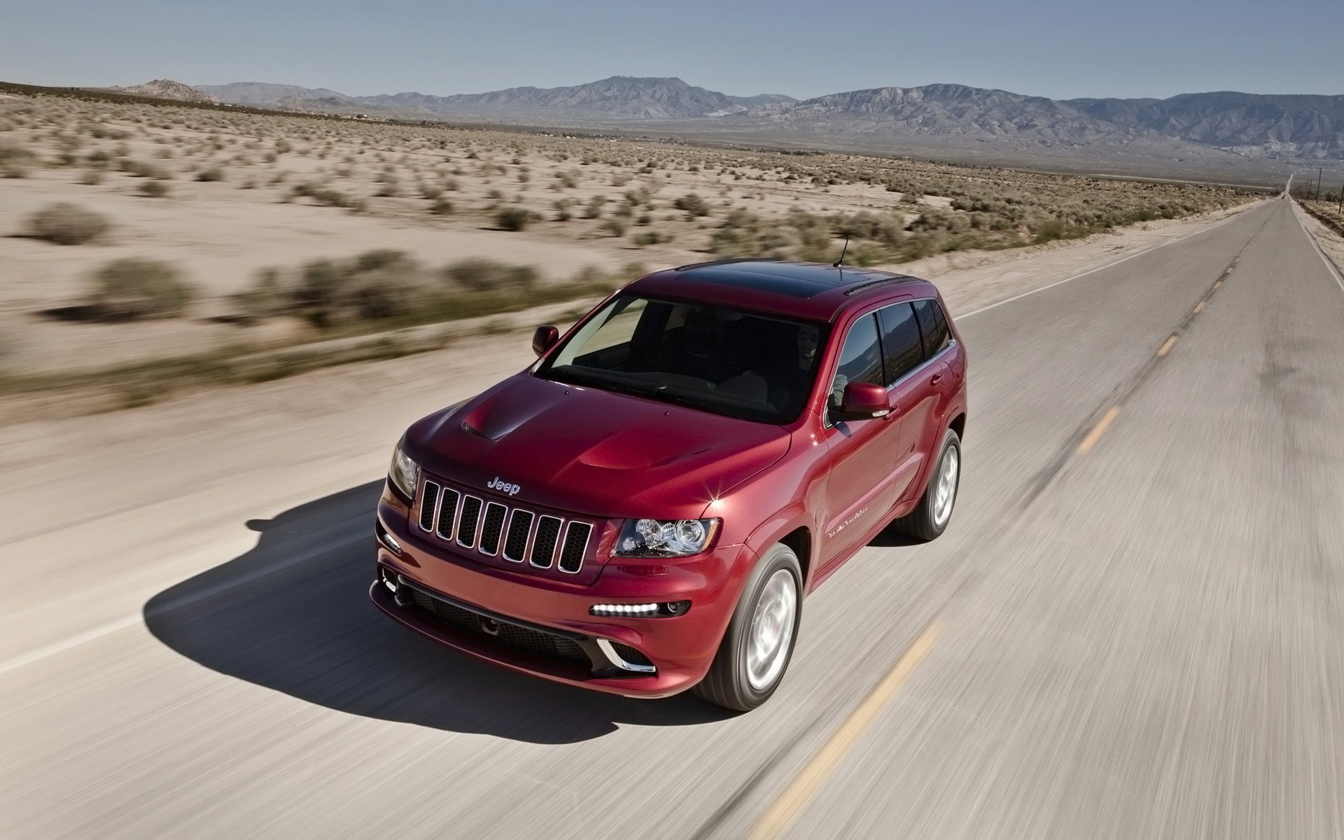 HD desktop wallpaper featuring a red Jeep SUV driving on a desert highway with mountains in the background.