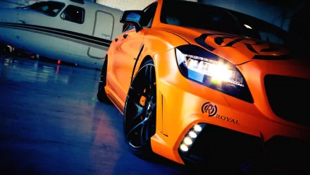 HD desktop wallpaper featuring a vibrant orange Mercedes-Benz tuned vehicle with sleek black wheels, highlighted in a stylish indoor setting near a private jet.