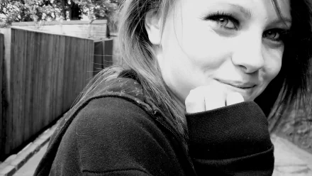 Black-and-white HD PC desktop wallpaper: close-up of a smiling young woman in a hoodie, hand to chin, outdoors beside a wooden fence, soft high-contrast portrait.