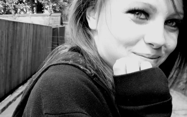Black-and-white HD PC desktop wallpaper: close-up of a smiling young woman in a hoodie, hand to chin, outdoors beside a wooden fence, soft high-contrast portrait.