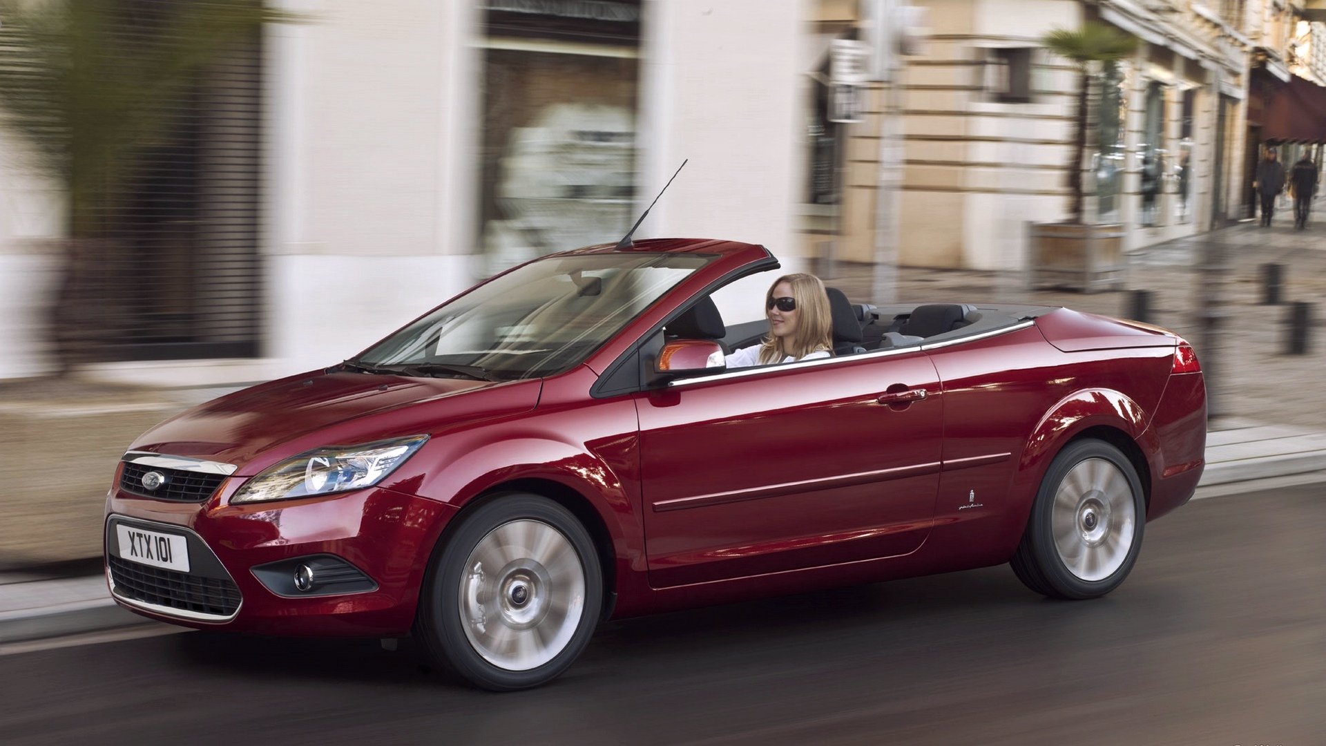 Red Ford Focus convertible cruising past city buildings, HD PC desktop wallpaper background.