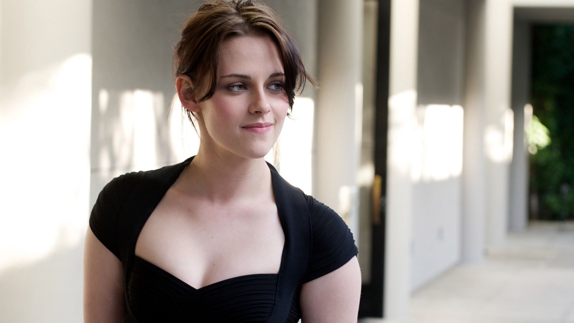 HD desktop wallpaper of American actress Kristen Stewart, looking to the side while standing in a well-lit hallway, wearing a black outfit.