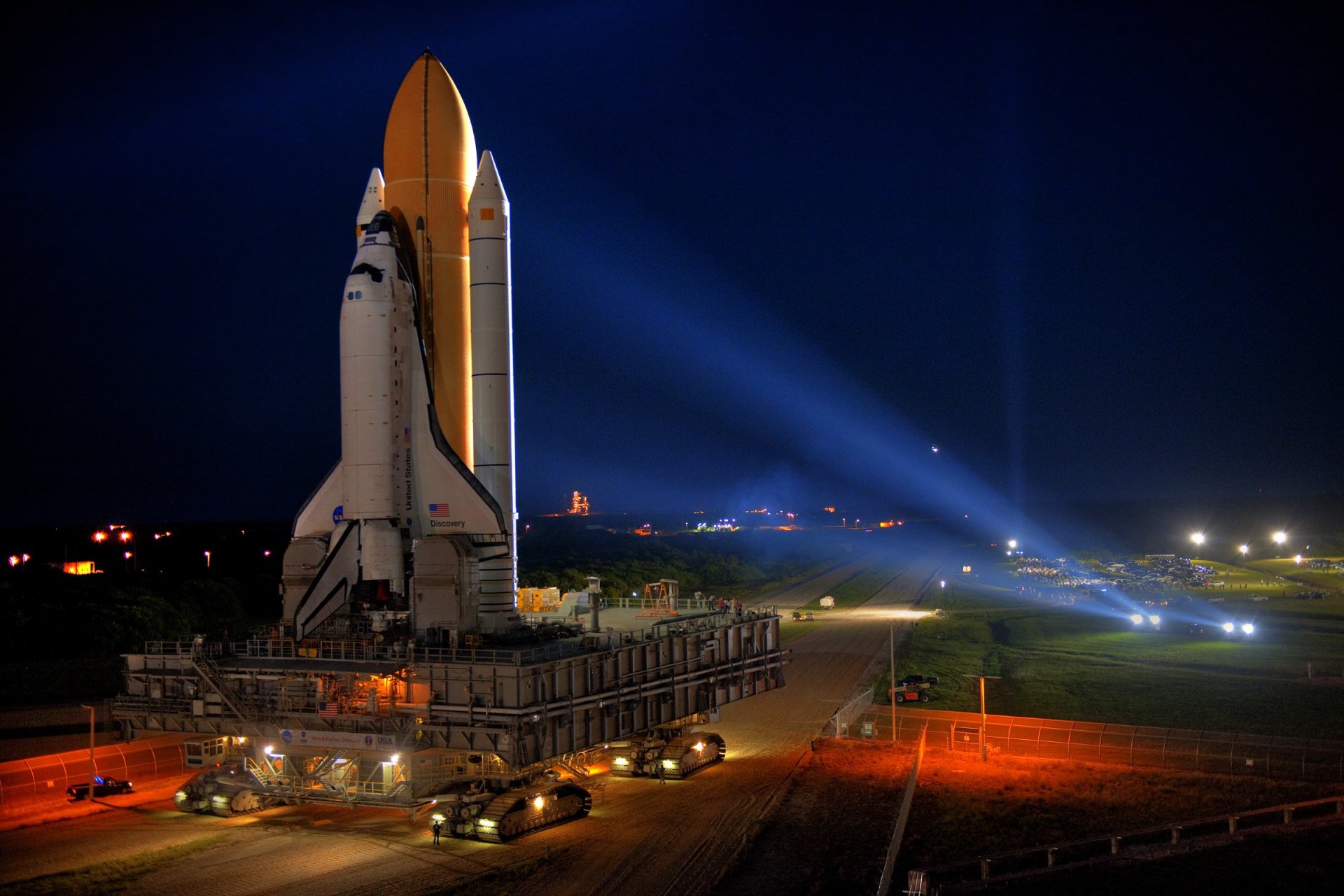 Space Shuttle Discovery stands on a launching pad at night, illuminated by bright spotlights, ready for its NASA mission.