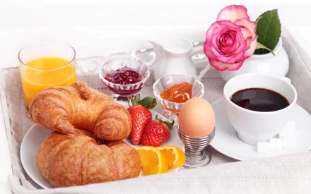A still life HD desktop wallpaper featuring a breakfast tray with croissants, strawberries, jams, a soft-boiled egg, coffee, orange juice, and a pink rose.
