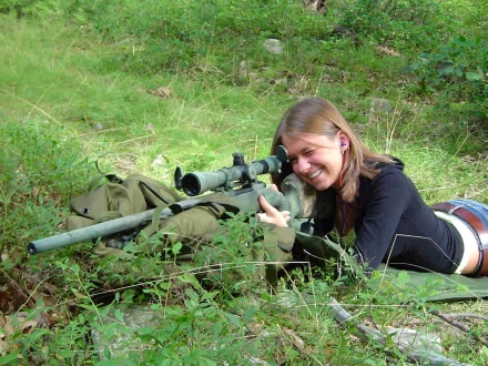 A woman lies in a grassy field, aiming a sniper rifle with a smile. This HD PC desktop wallpaper captures the theme of Girls & Guns in an engaging outdoor setting.