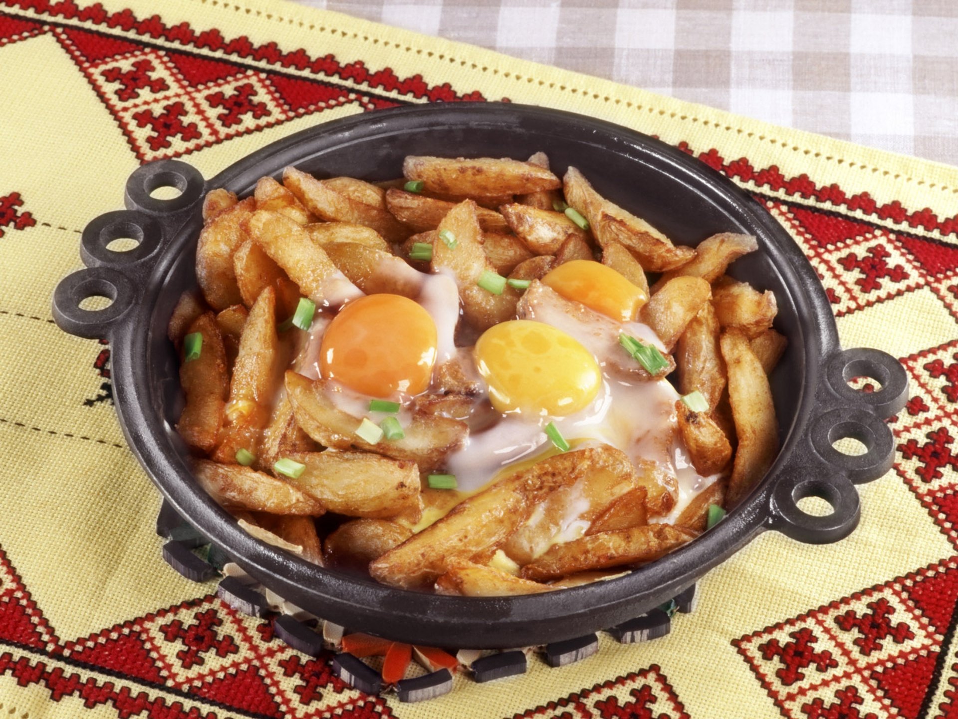 HD PC desktop wallpaper showing a hearty meal of fried potatoes topped with two sunny-side-up eggs, served in a rustic black pan on a patterned tablecloth.