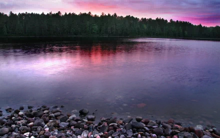HD desktop wallpaper of a tranquil river landscape with pebbles along the shore, calm water reflecting trees, and a vibrant pink and purple sunset sky.