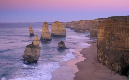 View of The Twelve Apostles rock formations along Australia's coastline with sandy beach, ocean waves, and cliffs under a pastel sunset sky.