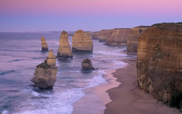View of The Twelve Apostles rock formations along Australia's coastline with sandy beach, ocean waves, and cliffs under a pastel sunset sky.