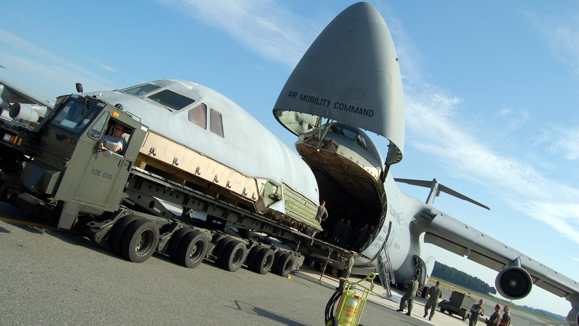 Military Lockheed C-5 Galaxy aircraft with nose open, loading cargo onto a large truck, captured in an HD PC desktop wallpaper background.