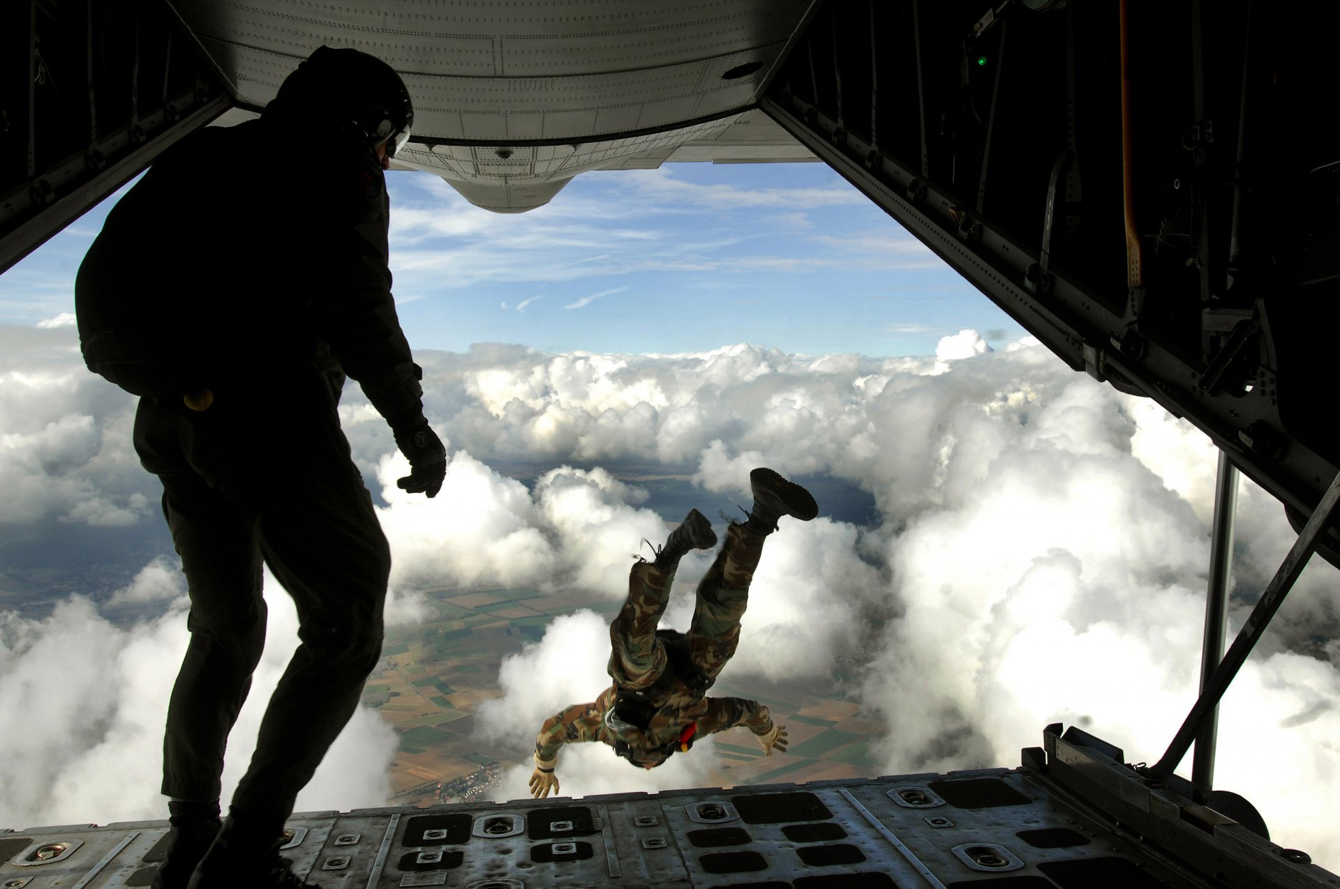 A paratrooper in military gear jumps from an aircraft, silhouetted against a backdrop of clouds, capturing the intensity and bravery of soldiers in action. HD desktop wallpaper.
