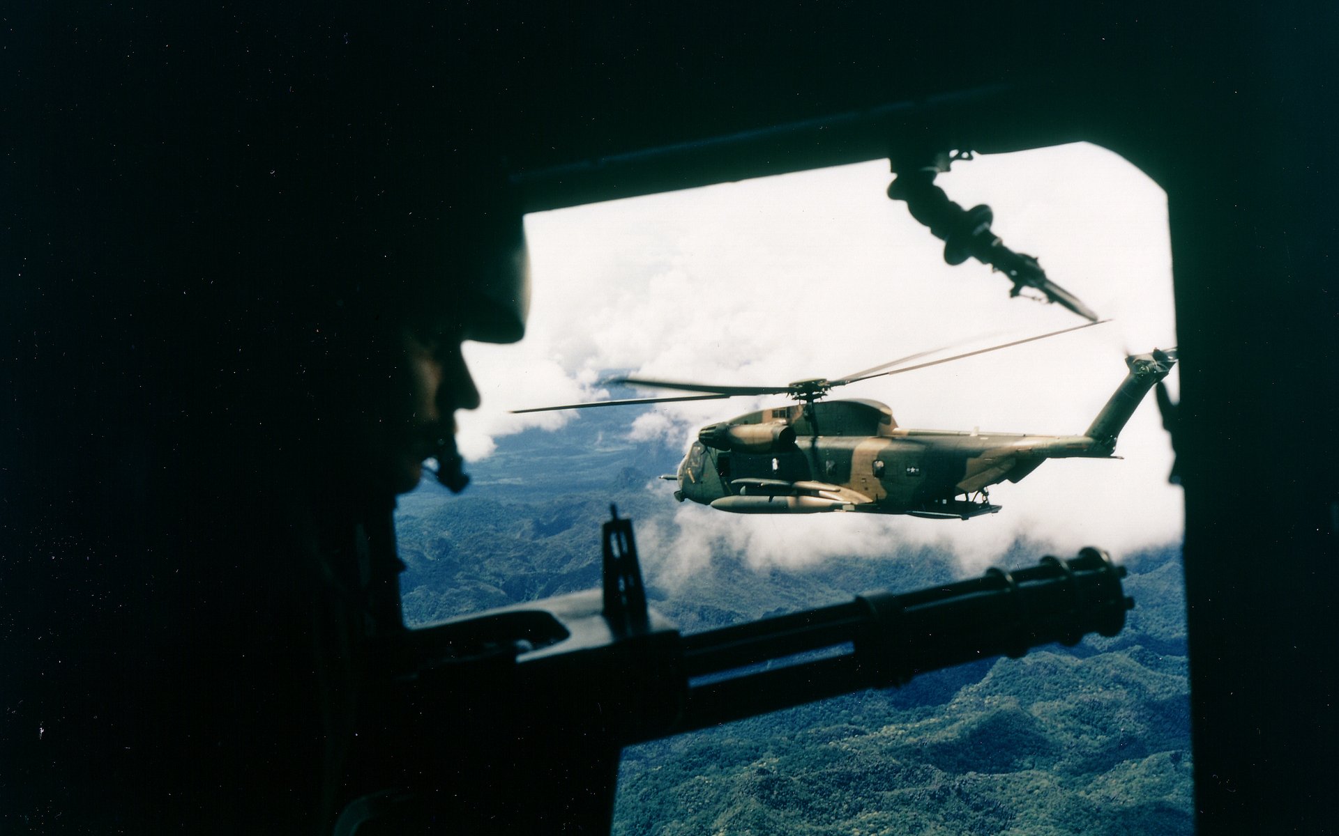 Silhouetted crew view from a transport door framing a Sikorsky MH-53 helicopter over cloud-covered jungle — military HD PC desktop wallpaper/background.
