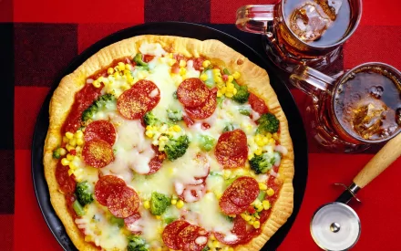 HD desktop wallpaper featuring a colorful pizza topped with broccoli, salami, and corn, accompanied by two glasses of Coca Cola on a red checkered tablecloth.