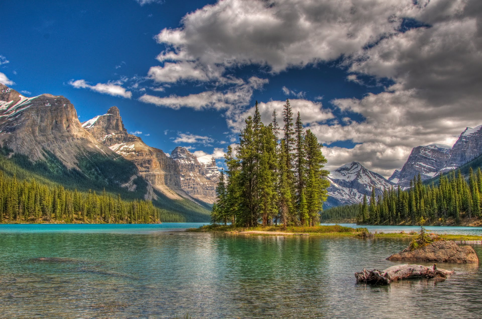 HD PC desktop wallpaper featuring a serene nature landscape with a small island of pine trees in a clear blue lake, surrounded by rugged mountains under a partly cloudy sky.