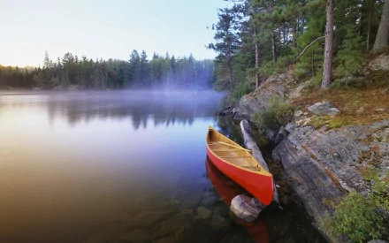 A red canoe rests along a rocky shore of a foggy river or lake, surrounded by dense forest, captured in a serene HD PC desktop wallpaper.