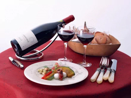 A lunch setting on a red tablecloth with a plate of vegetables, two glasses of red wine, a wine bottle in a stand, and a basket of bread, captured in HD desktop wallpaper quality.
