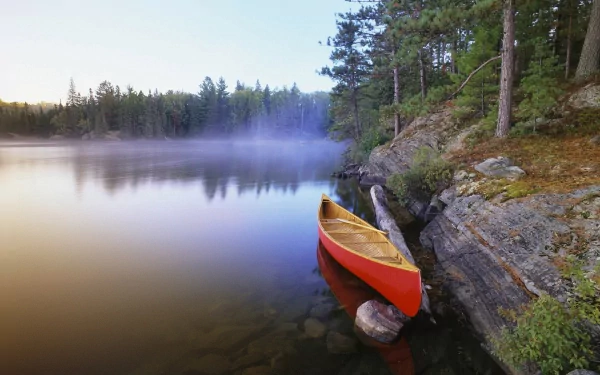 A red canoe rests along a rocky shore of a foggy river or lake, surrounded by dense forest, captured in a serene HD PC desktop wallpaper.