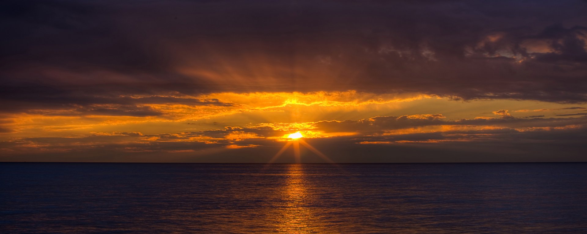 HD desktop wallpaper of a vibrant ocean sunset with golden rays breaking through dark clouds over calm water, showcasing stunning natural beauty.