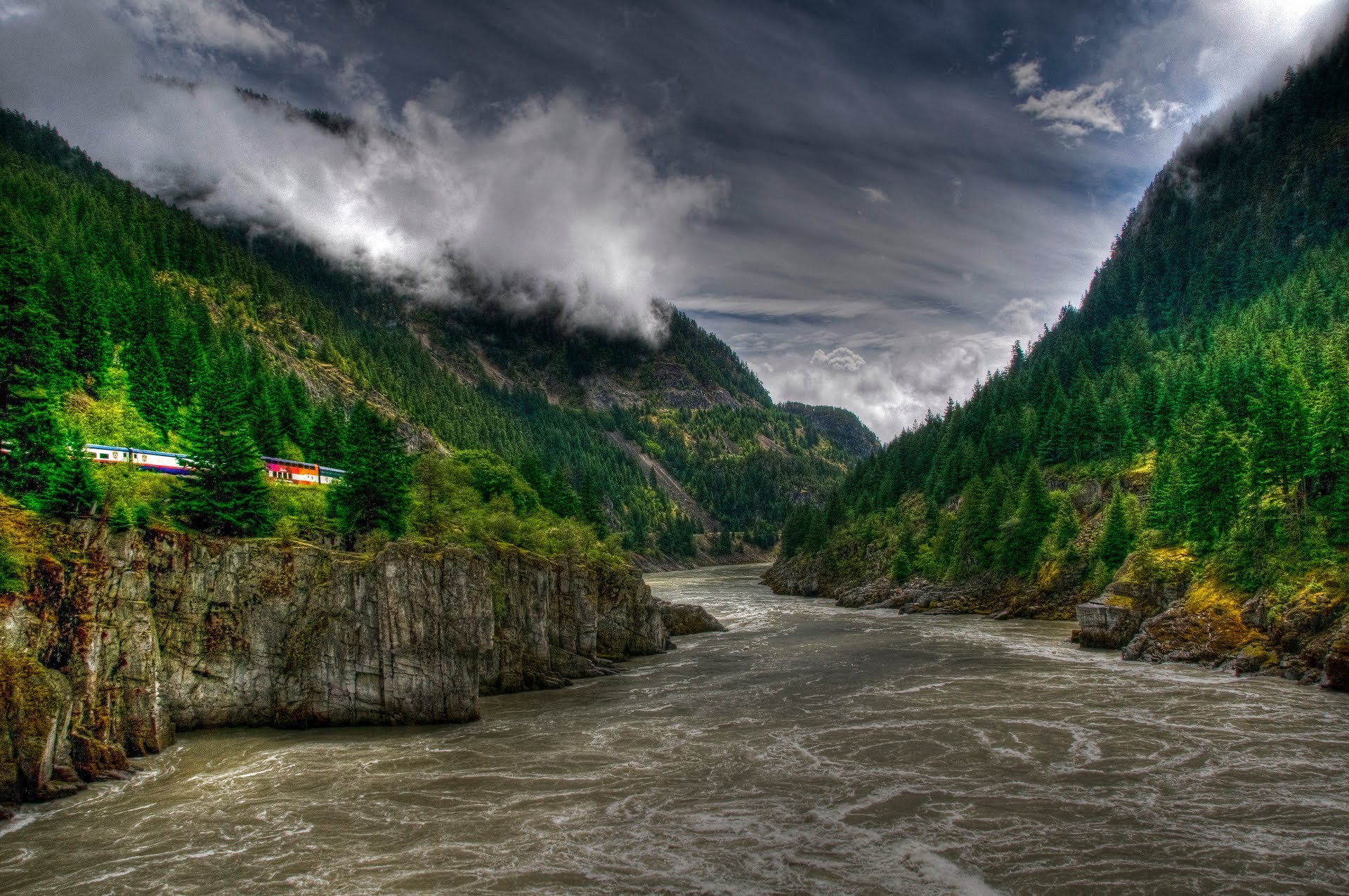 Dramatic landscape of the Fraser River flowing through Hell's Gate, British Columbia, surrounded by lush forests and rugged cliffs under a moody sky.