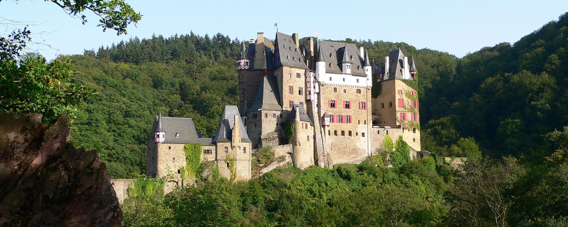 HD PC desktop wallpaper of a man-made medieval castle perched on a forested hill, sunlit stone towers, turrets and battlements against a clear blue sky.