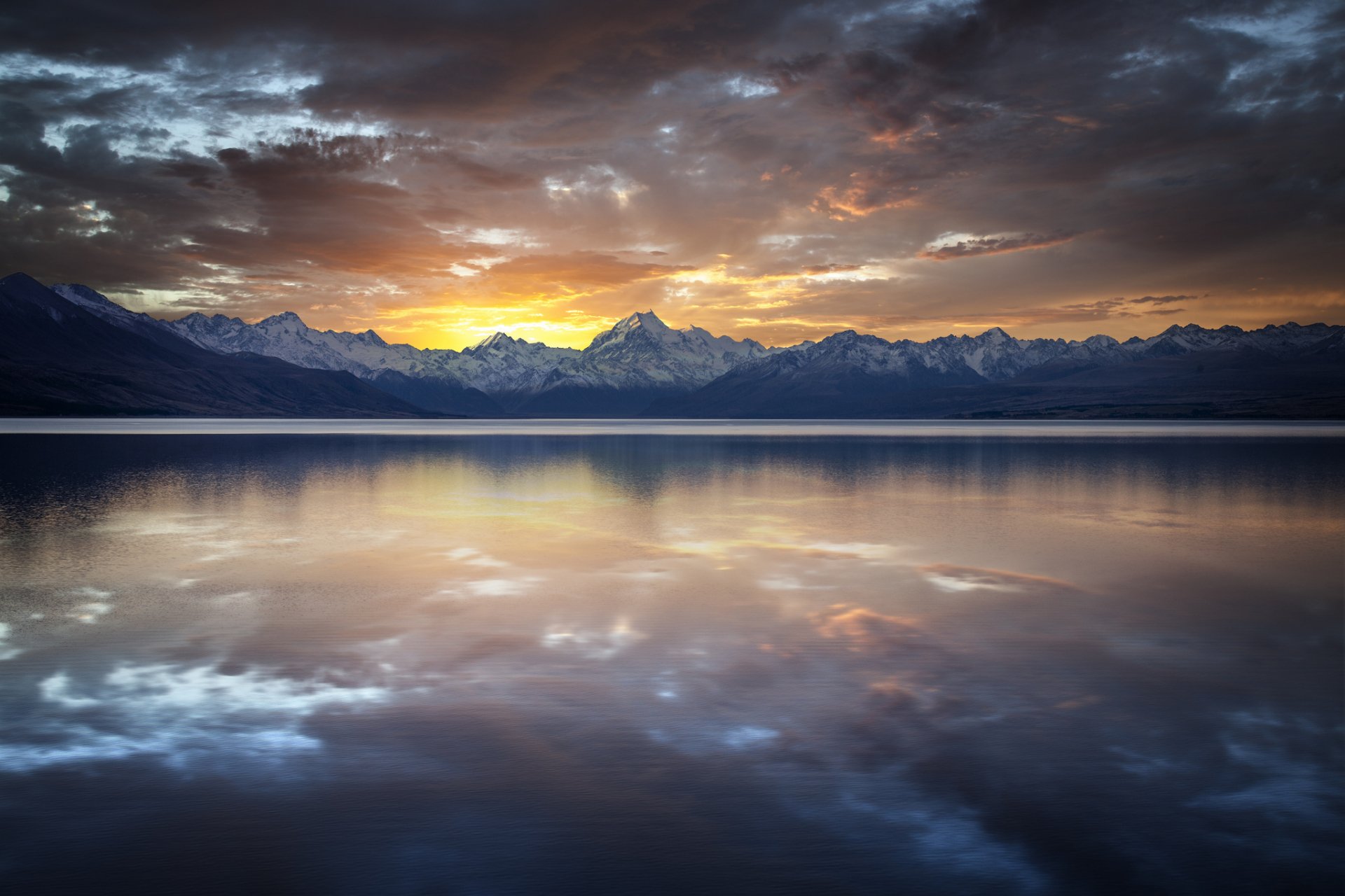 A tranquil lake reflects the majestic mountains under a dramatic sky at sunset, with vibrant colors illuminating the clouds and water, capturing the beauty of nature.