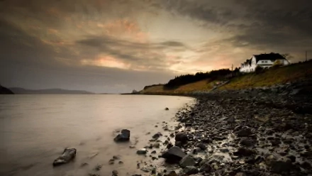 Photography of a rocky shoreline at dusk with a house on a hill, serving as an HD PC desktop wallpaper and background capturing a serene coastal place.