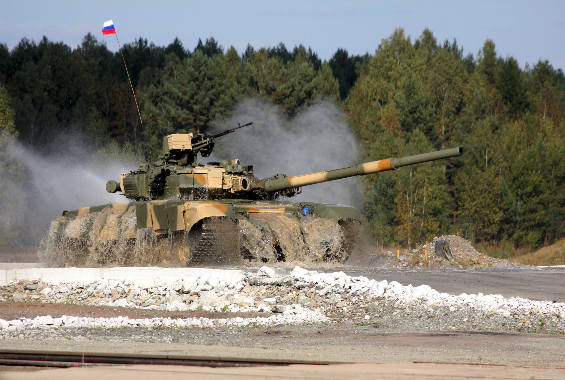 A 4K Ultra HD desktop wallpaper showcasing a T-90 military tank moving on a dirt road with dust clouds, set against a forest backdrop under a clear sky.