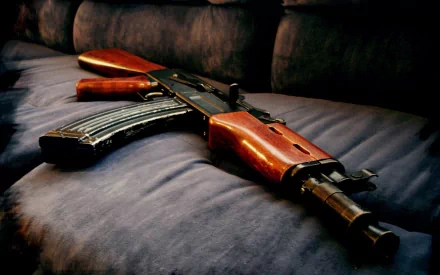 HD desktop wallpaper featuring a man-made AKM assault rifle with a wooden stock and grip, placed on a dark fabric surface. The image highlights the weapon's detailed craftsmanship against a soft, blurred background.