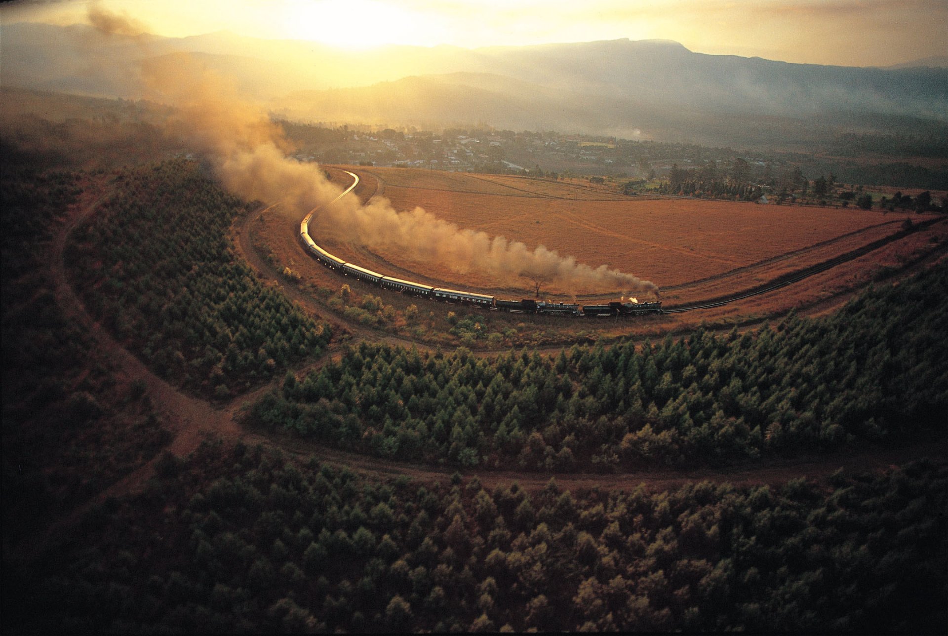 A breathtaking HD desktop wallpaper showcasing an aerial view of a train curving through a vast rural landscape with the sun setting in the background, casting a warm, golden glow over the scenery.