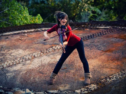 Lindsey Stirling plays the violin joyfully on a textured, rust-colored surface, surrounded by lush greenery, captured in an HD desktop wallpaper.