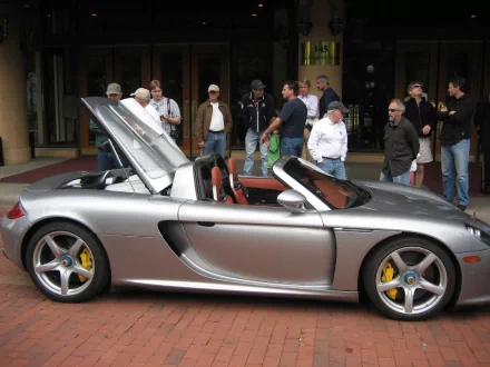 A sleek silver Porsche Carrera GT with its hood open, surrounded by a crowd of onlookers, set against a vibrant backdrop. An eye-catching HD desktop wallpaper.