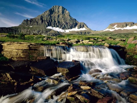 HD desktop wallpaper of Glacier National Park in Montana, showcasing a cascading waterfall in the foreground with a majestic mountain in the background, surrounded by lush greenery and patches of snow.