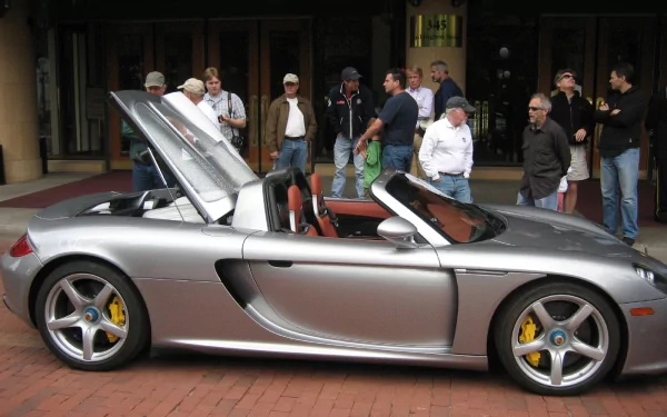 A sleek silver Porsche Carrera GT with its hood open, surrounded by a crowd of onlookers, set against a vibrant backdrop. An eye-catching HD desktop wallpaper.