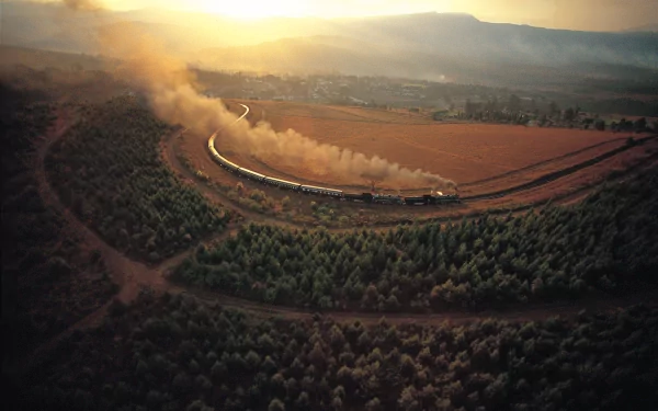 A breathtaking HD desktop wallpaper showcasing an aerial view of a train curving through a vast rural landscape with the sun setting in the background, casting a warm, golden glow over the scenery.