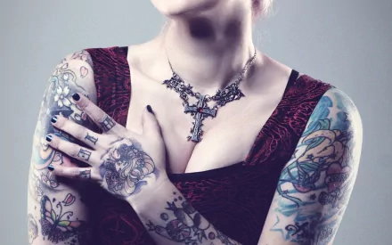 A woman with numerous colorful tattoos on her arms and hands poses with confidence, wearing a cross necklace. The image is a high-definition desktop wallpaper and background.