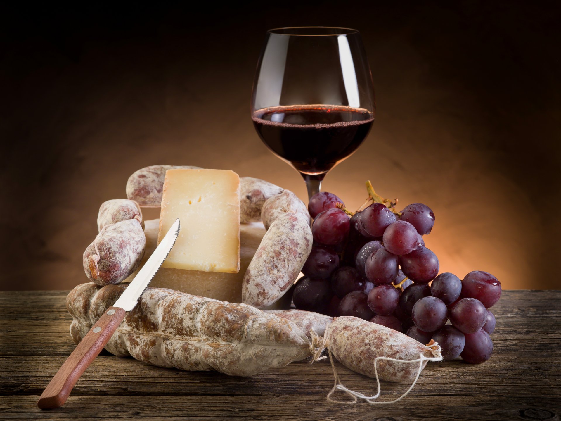 A still life composition featuring cured sausages, a wedge of cheese, fresh grapes, and a glass of red wine, presented in rich colors for a 4K Ultra HD desktop background.