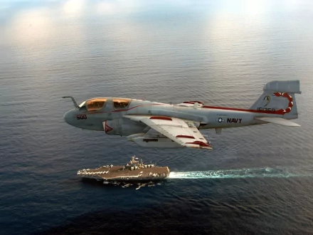 A Northrop Grumman EA-6B Prowler flies over a naval aircraft carrier in the ocean, captured in a high-definition military-themed desktop wallpaper and background.