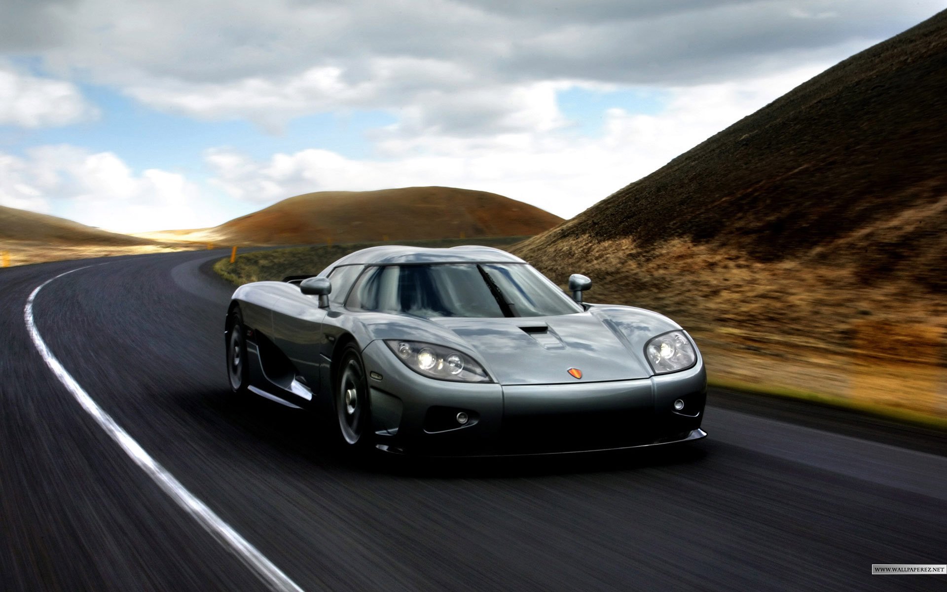 HD PC desktop wallpaper featuring a sleek silver Koenigsegg sports car driving on a winding road through a hilly landscape under a cloudy sky.
