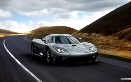 HD PC desktop wallpaper featuring a sleek silver Koenigsegg sports car driving on a winding road through a hilly landscape under a cloudy sky.