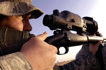 A close-up HD desktop wallpaper of a military sniper in camouflage aiming through a high-powered rifle scope against a clear sky backdrop.