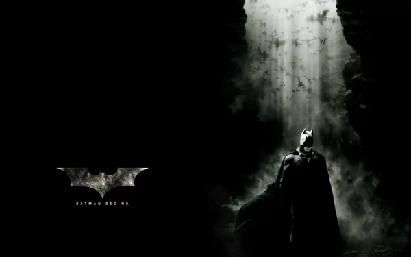 HD desktop wallpaper featuring Batman from the movie Batman Begins. The image shows Batman standing in a dark cave with light shining down and bats flying overhead. The Batman logo is on the left.