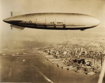 An aerial view of a U.S. Navy airship gliding over a coastal city, captured in a historical sepia tone. This HD wallpaper captures the essence of military aviation.