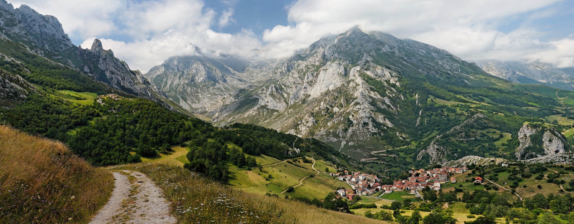 HD desktop wallpaper featuring a scenic landscape of a town nestled in green hills with a winding path, surrounded by towering mountains under a partly cloudy sky.