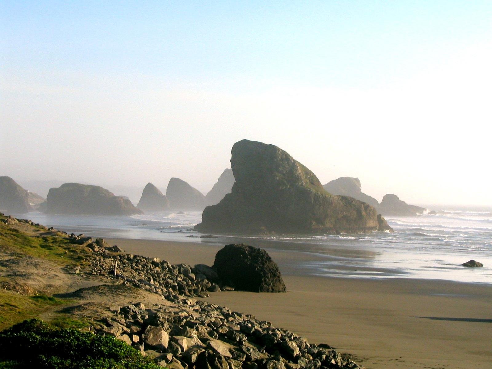 A serene beach with large rock formations rising from the ocean, framed by a soft mist and gentle waves. This HD wallpaper beautifully captures the tranquil essence of nature.